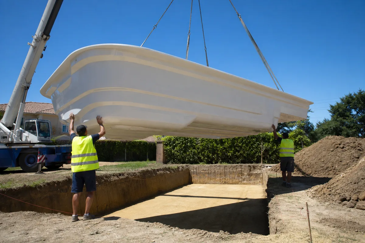 Chantier de pose d&rsquo;une coque polyester grutée au-dessus d&rsquo;une fouille de jardin en plein soleil