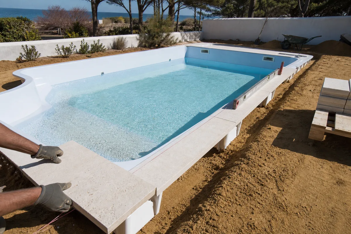 Coque de piscine posée sur lit de sable stabilisé avec margelles claires en cours de pose